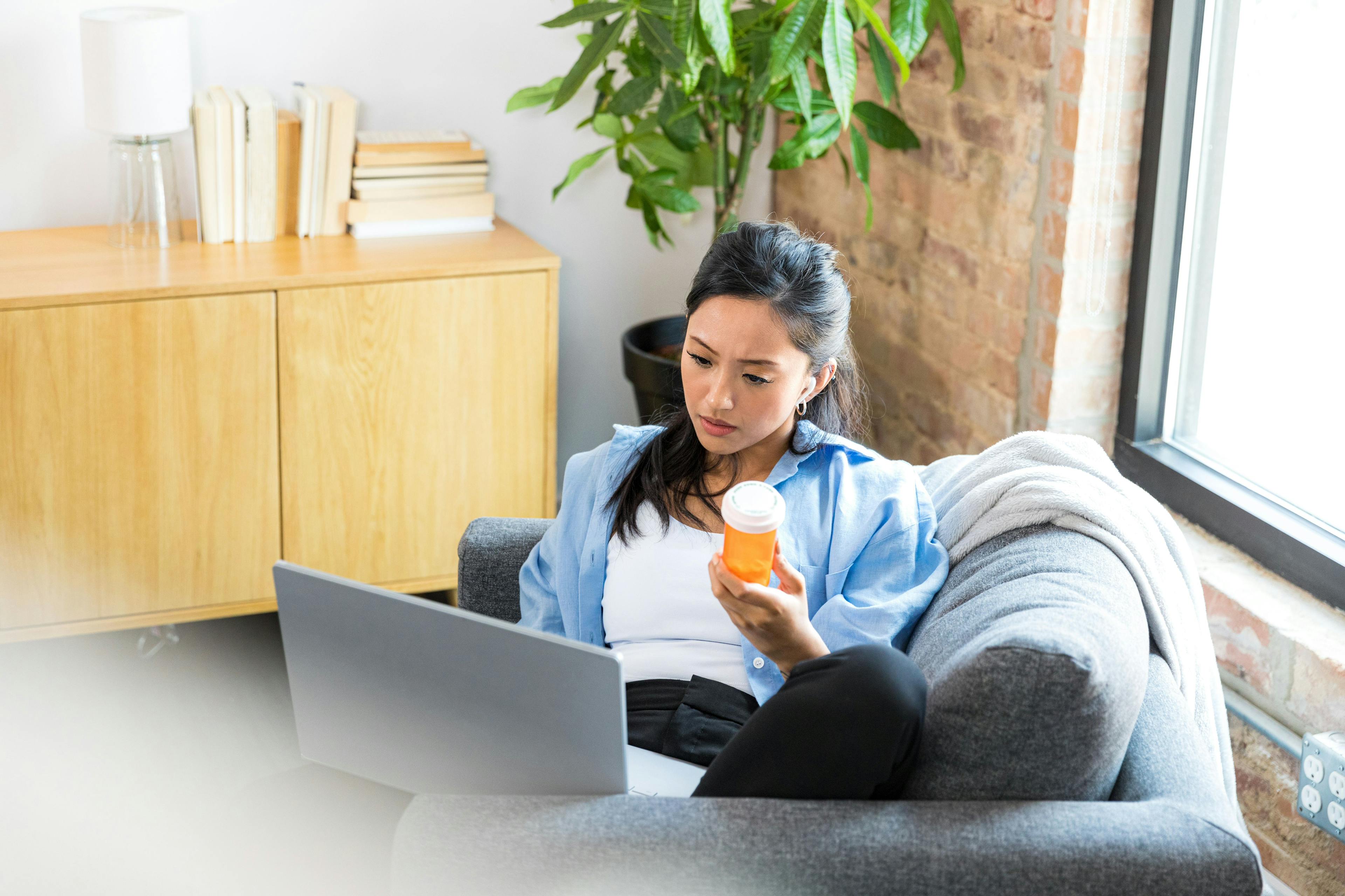 A woman sits on her couch holding a pill bottle in one hand and her laptop in the other.