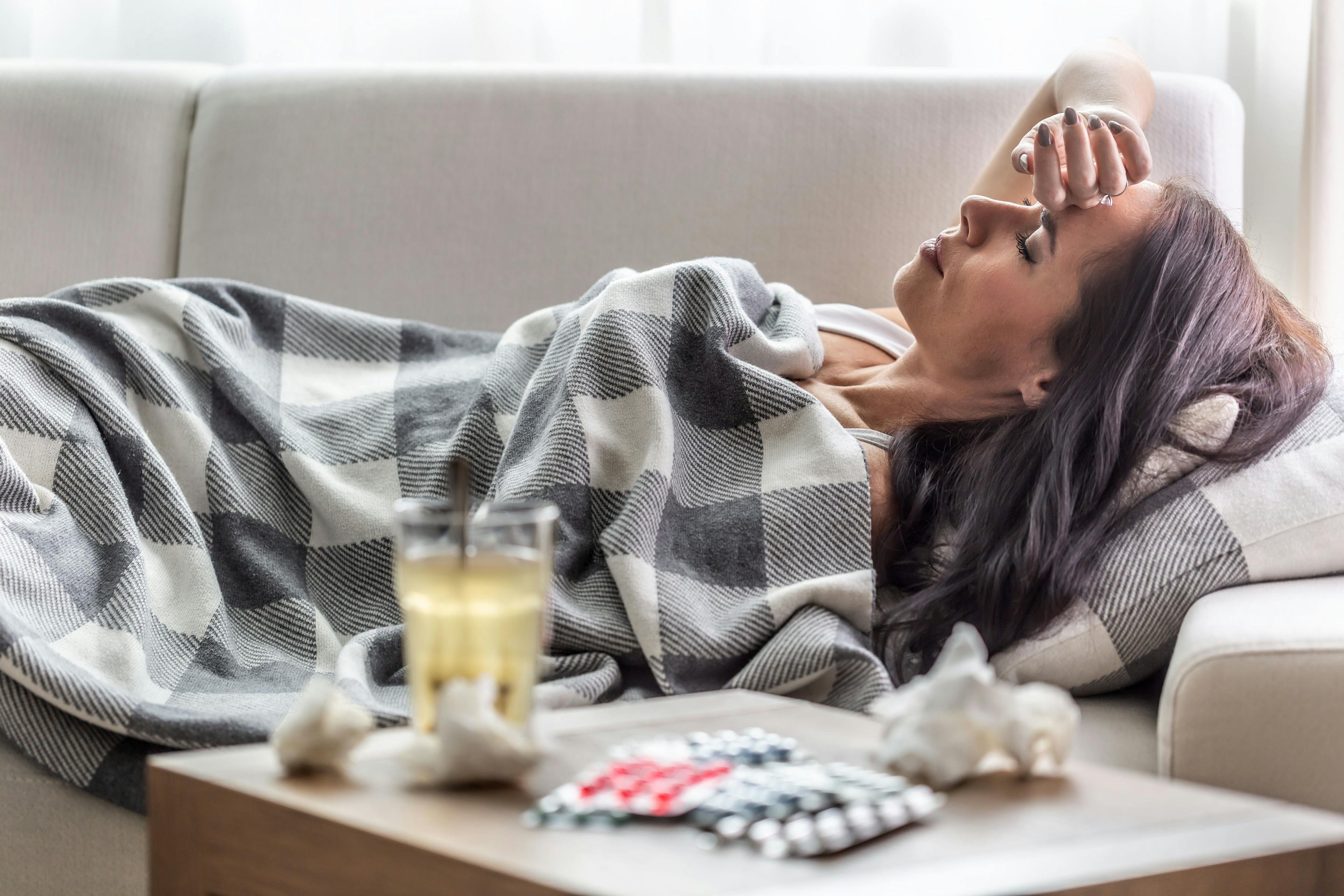 A young, sick woman is laying on her couch with a checked blanket over her and tissues, pills and a drink next to her.